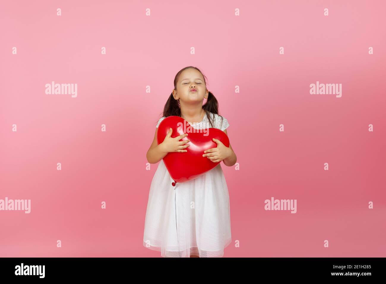 Ein Mädchen mit geschlossenen Augen in einem weißen Kleid hält einen roten Ballon in Form eines Herzens und bläst einen Kuss, isoliert auf einem rosa Hintergrund Stockfoto