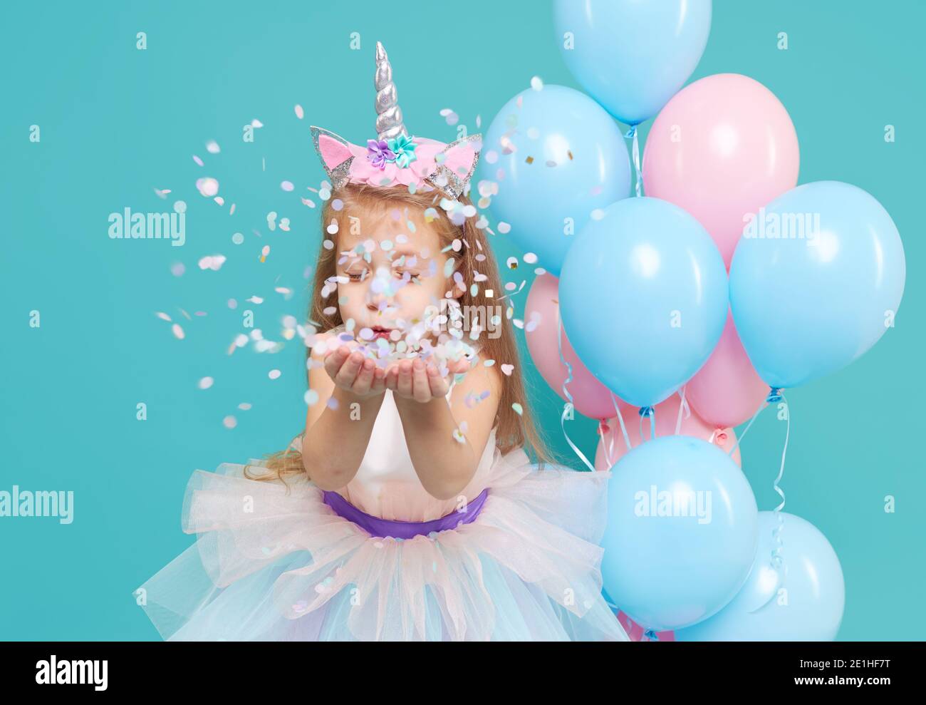 Einhorn-Mädchen. Fröhliche Kind Mädchen in eleganten Tüllkleid und Einhorn Stirnband feiert Geburtstagsfeier und weht bunten Konfetti auf cyan Rücken Stockfoto