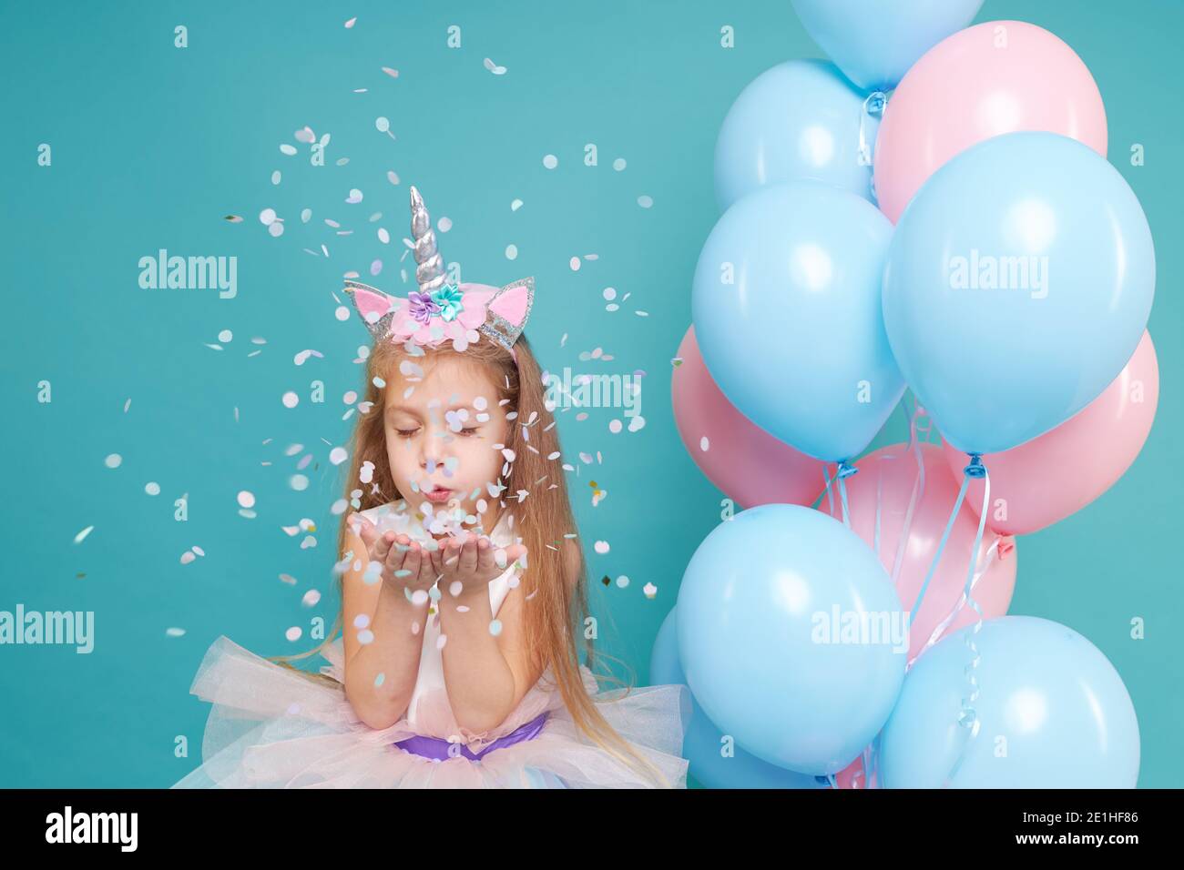 Einhorn-Mädchen. Fröhliche Kind Mädchen in eleganten Tüllkleid und Einhorn Stirnband feiert Geburtstagsfeier und weht bunten Konfetti auf cyan Rücken Stockfoto