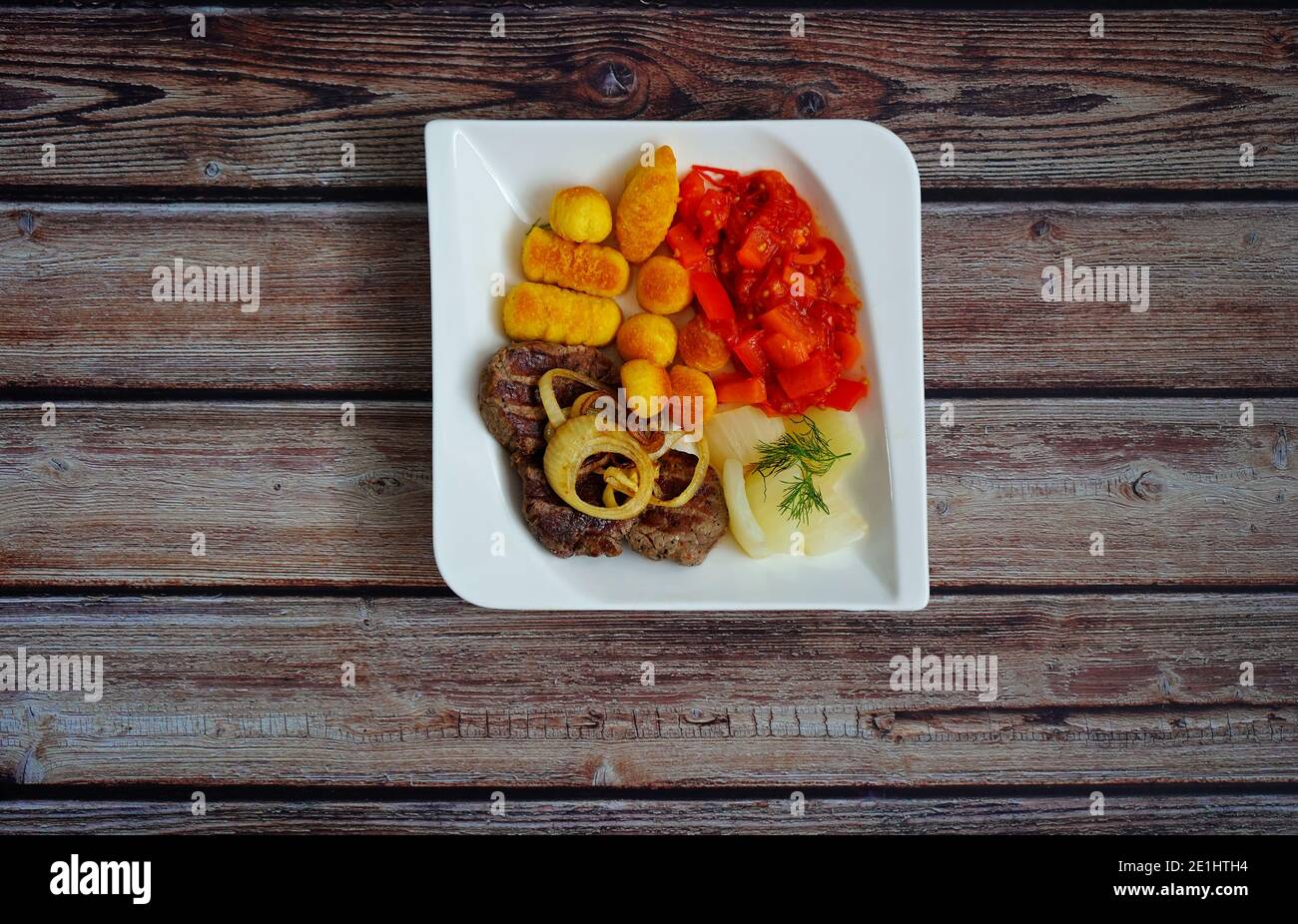 Rindersteak mit Zwiebelringen, Kroketten und Gemüse (Fenchel und Paprika) auf einem weißen quadratischen Teller mit dunklem Holzhintergrund. Deutsche Hausmannskost. Stockfoto