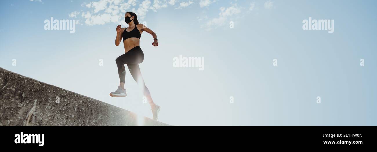 Low-Angle-Aufnahme einer Frau beim Laufen im Freien mit Sonne scheint von hinten. Weiblich in Sportbekleidung und schützende Gesichtsmaske Ausübung Outdoo Stockfoto