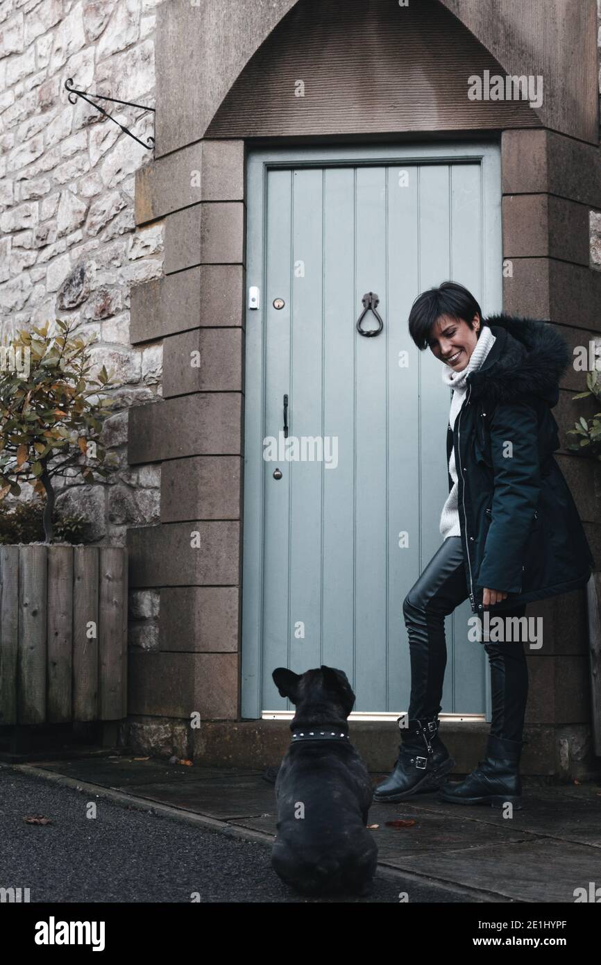 Fröhliche Frau in Winterkleidung, die nach Hause kommt und lächelt an Ihr Hund, während der Hund auf sie zurückblickt Stockfoto