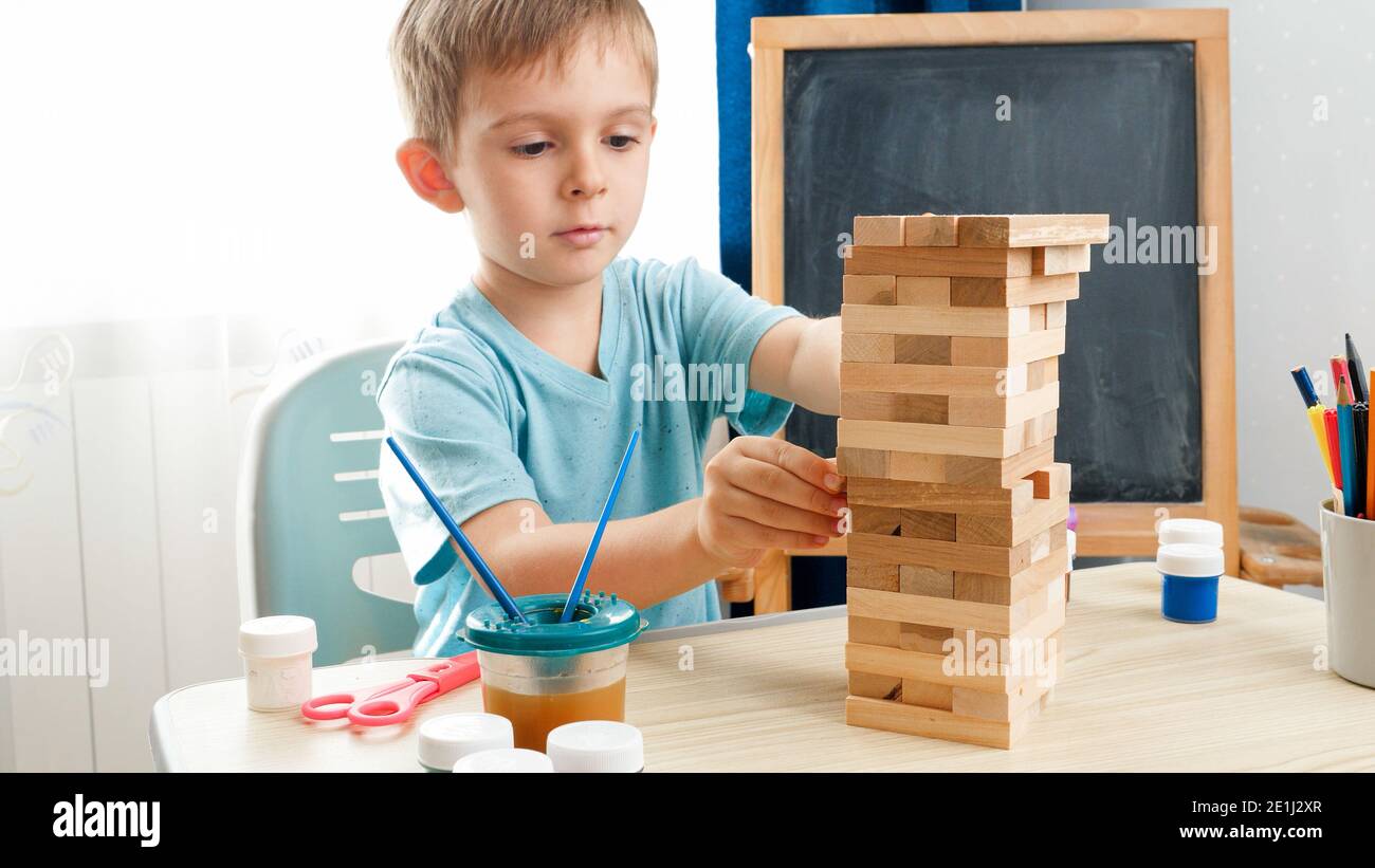 Intelligenter Junge, der Holzblöcke aus dem hohen Turm aus Ziegelsteinen zieht. Konzept von intelligenten Kindern und häuslicher Bildung während des Lockdown und der Selbstisolation. Stockfoto