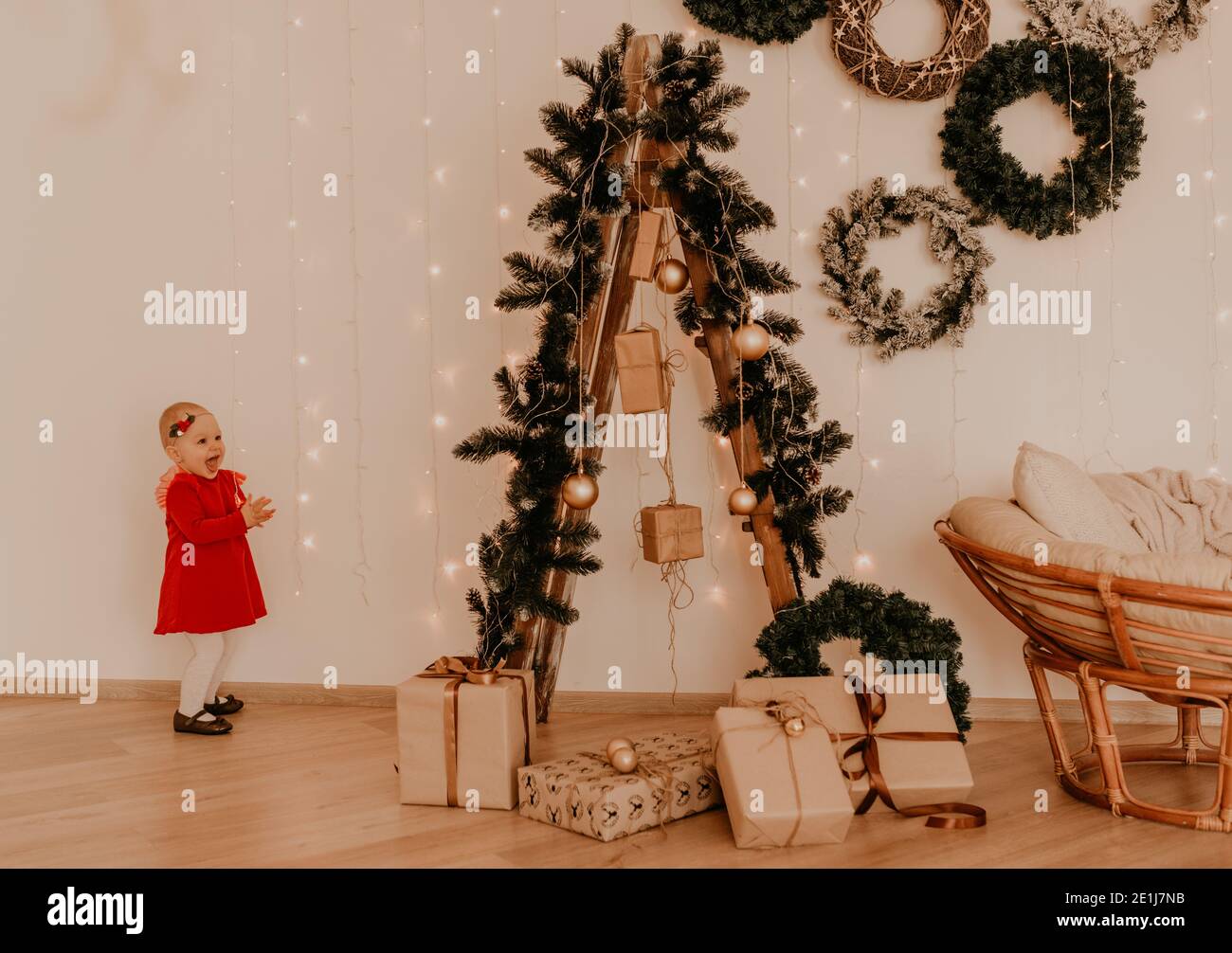 Kleines Mädchen in roten Kleid läuft Wohnung Stil Licht Minimalismus. Kind steht an der weißen Wand mit Girlanden Weihnachtsbaum mit eingewickelten Geschenken. Weihnachten. Stockfoto