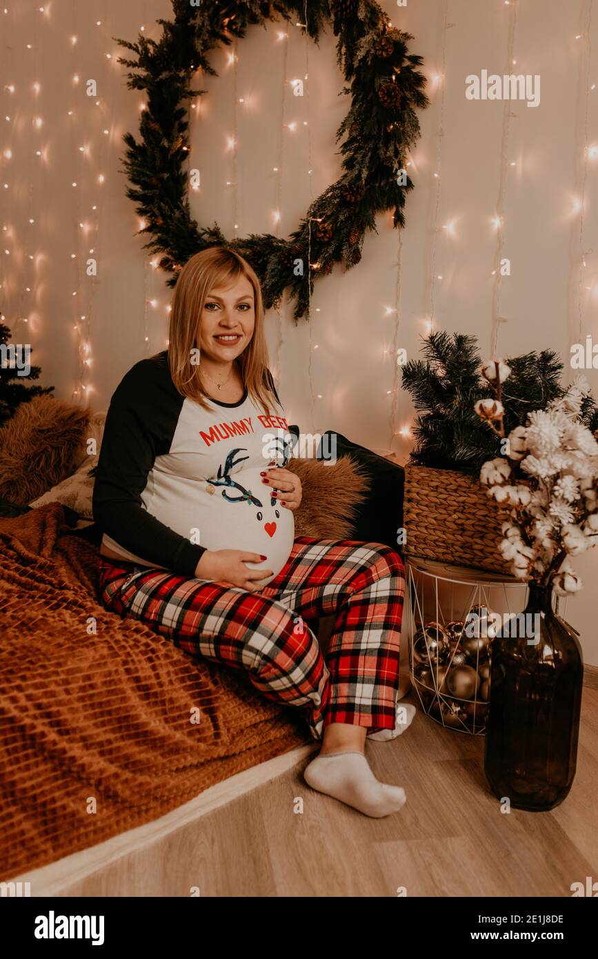 Schwangere Frau sitzt im Schlafanzug mit auf dem Bett im Schlafzimmer. Weihnachtsmorgen. Das Interieur des neuen Jahres. Valentinstag-Feier Stockfoto