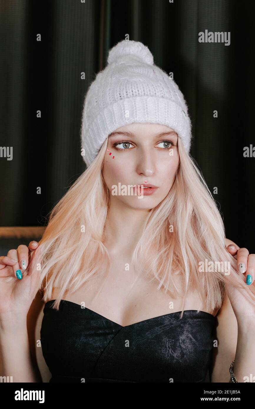 Hübsches blondes Mädchen trägt einen gestrickten warmen Hut mit Pompon und ein stilvolles dunkles trägerloses Kleid; sie zieht ihr Haar mit beiden Händen an die Seiten Stockfoto