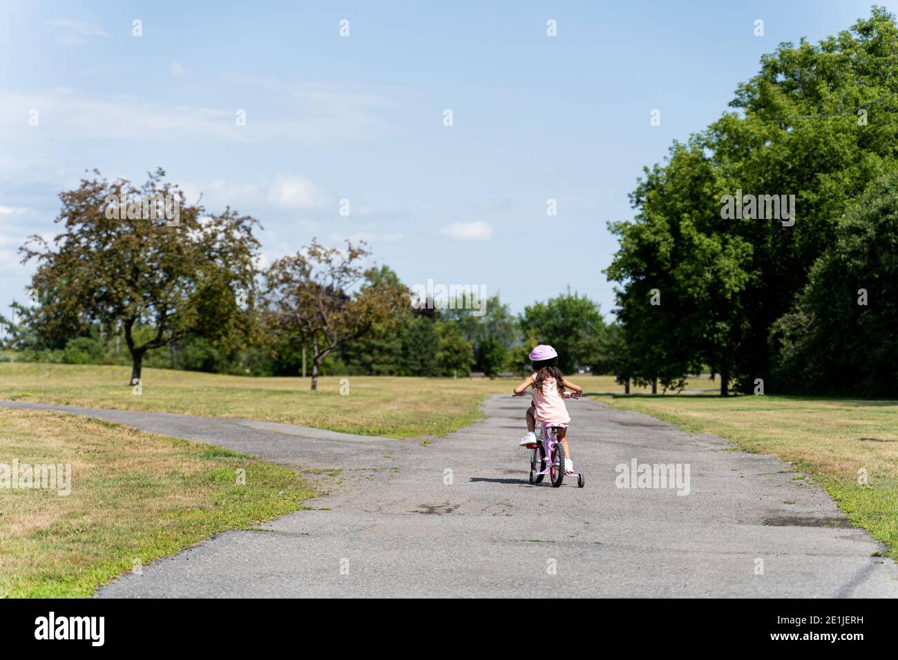 Junges Mädchen in rosa Kleid und Helm Reiten rosa Fahrrad auf einem Park Bürgersteig vor der Kreuzung, von hinten gesehen Stockfoto