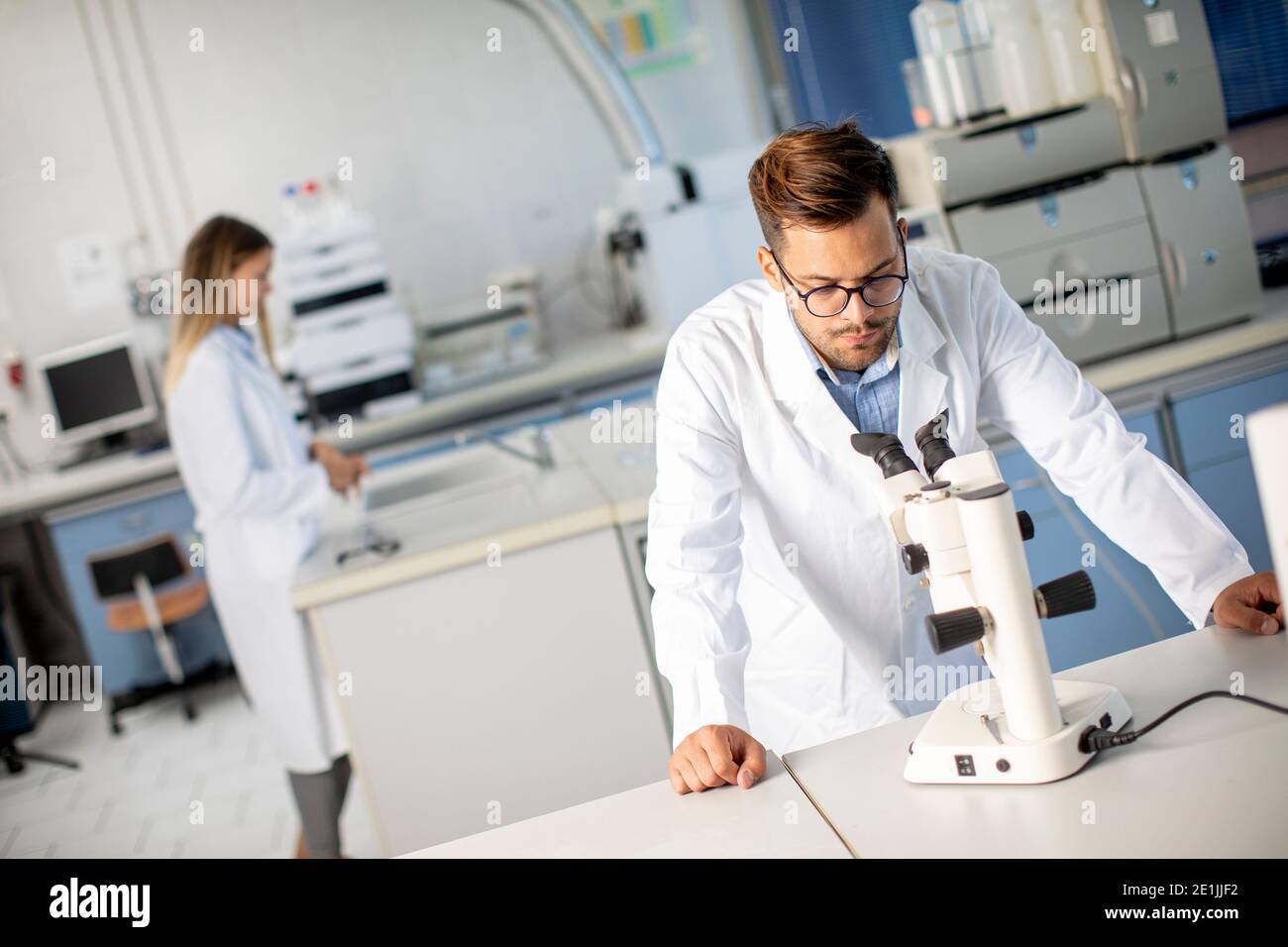 Hübscher junger Wissenschaftler im weißen Laborkittel, der mit binokular arbeitet Mikroskop im Labor der Materialwissenschaft Stockfoto