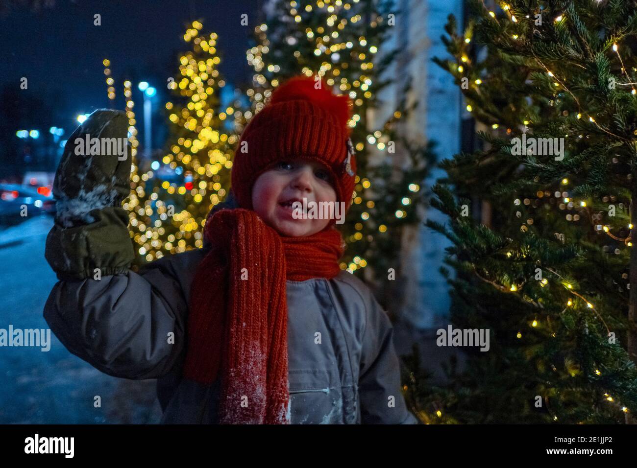 Glückliches Kind in warmer Oberbekleidung winkende Hand und Blick auf Kamera in der Nähe von Fichte mit leuchtenden Lichtern geschmückt stehen Abends Stadt während C Stockfoto