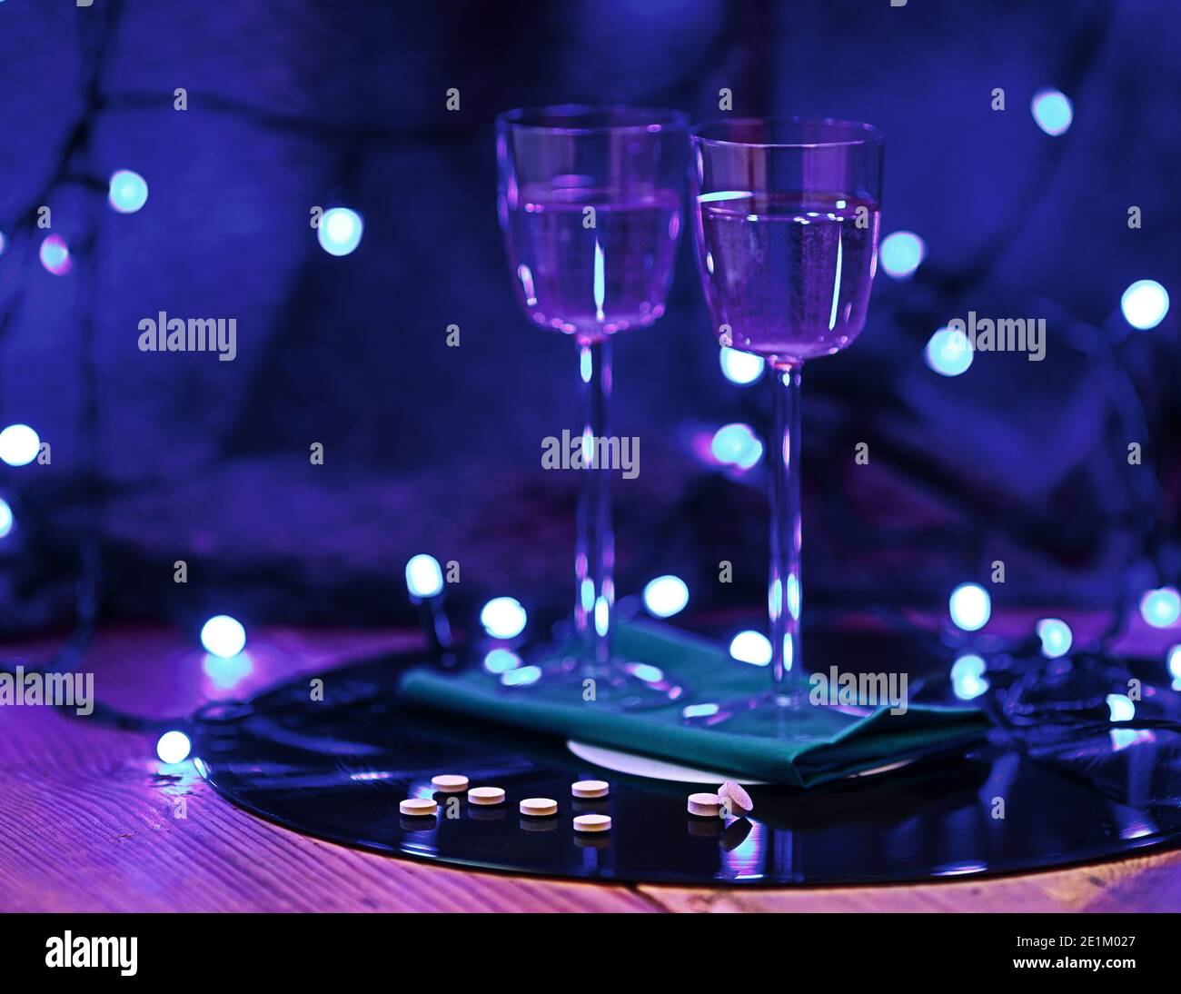 Selektiver Fokus Foto von Pillen vor dem Hintergrund von zwei Gläsern Champagner und einer Vinyl-Platte, umgeben von hellen Club Beleuchtung Stockfoto