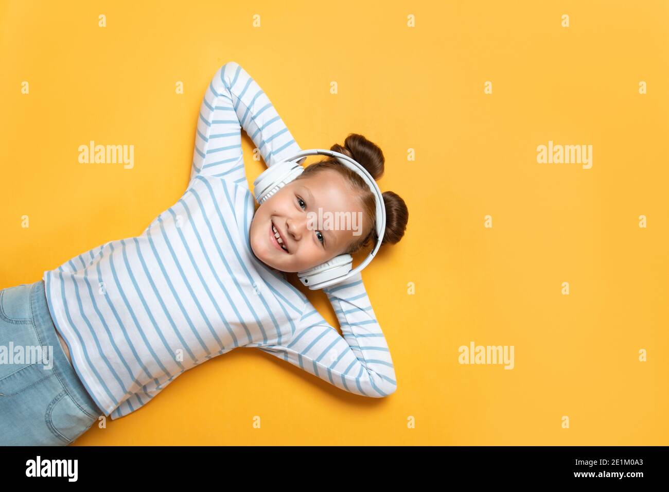 Fröhliche niedliche kleine Mädchen in guter Stimmung hören moderne Technologie Kopfhörer auf einem gelben Hintergrund. Das Kind liegt mit den Händen hinter seinem er Stockfoto