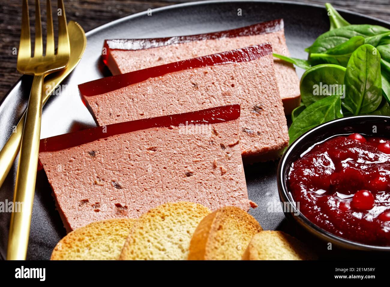 Französisches Gericht Entenleber Pastete mit Cranberry Marmelade auf einem schwarzen Teller serviert mit Cranberry-Sauce, Spinat und Brioche Croutons, mit goldenem c Stockfoto
