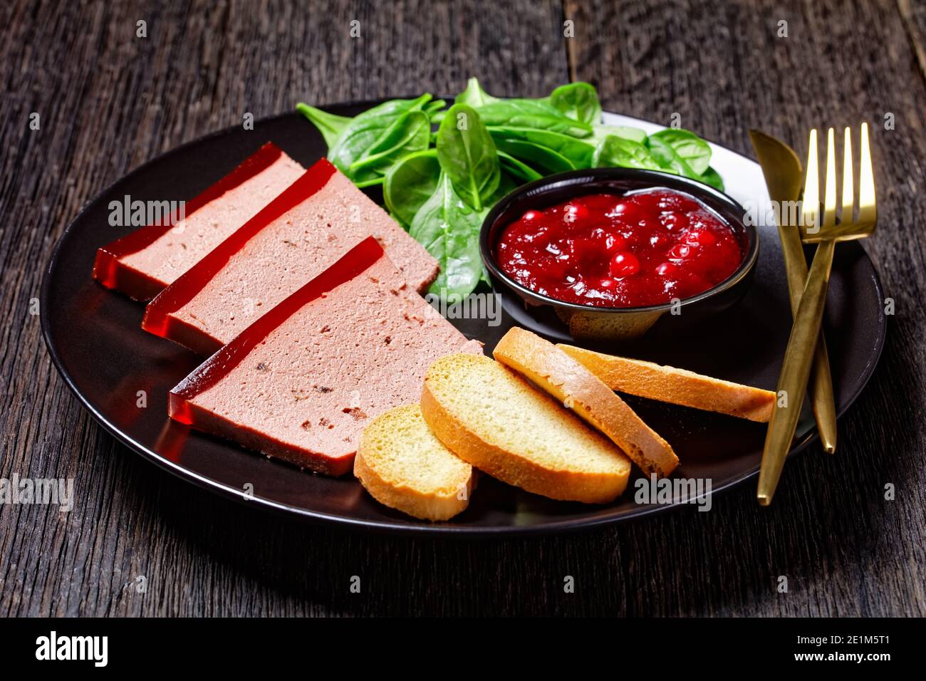 Französisches Gericht Entenleber Pastete mit Cranberry Marmelade auf einem schwarzen Teller serviert mit Cranberry-Sauce, Spinat und Brioche Croutons, mit goldenem c Stockfoto
