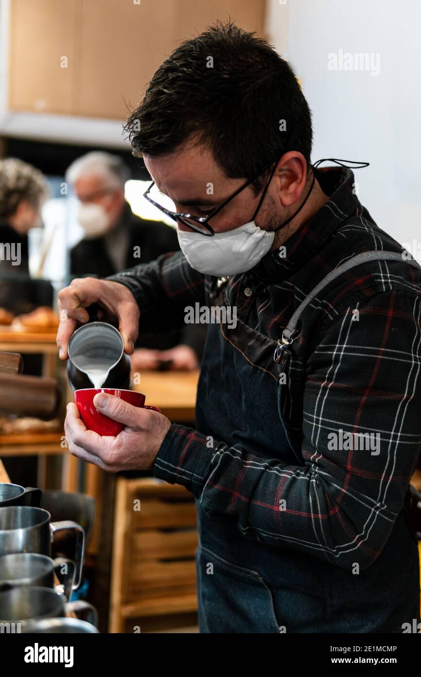 Kaukasischer Barista mit Maske gießt Milch in eine rote Kaffeetasse in einem Coffee Shop, vor der Kaffeemaschine mit zwei unfokussierten Kunden im Rücken Stockfoto