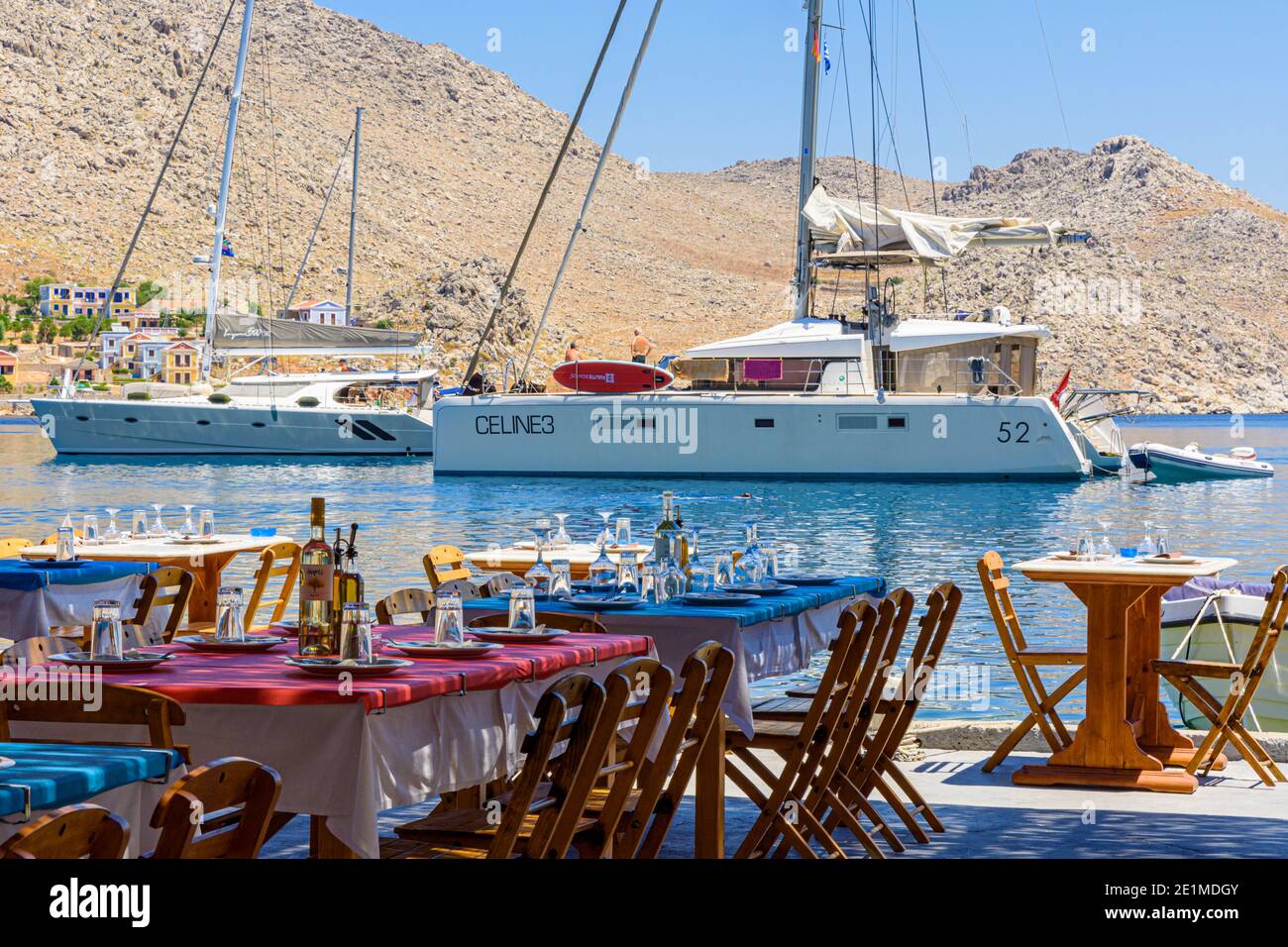 Taverna Tische und Stühle am Wasser mit Blick auf verankerte Boote in Pedi Bay, Symi Island, Dodekanes, Griechenland Stockfoto