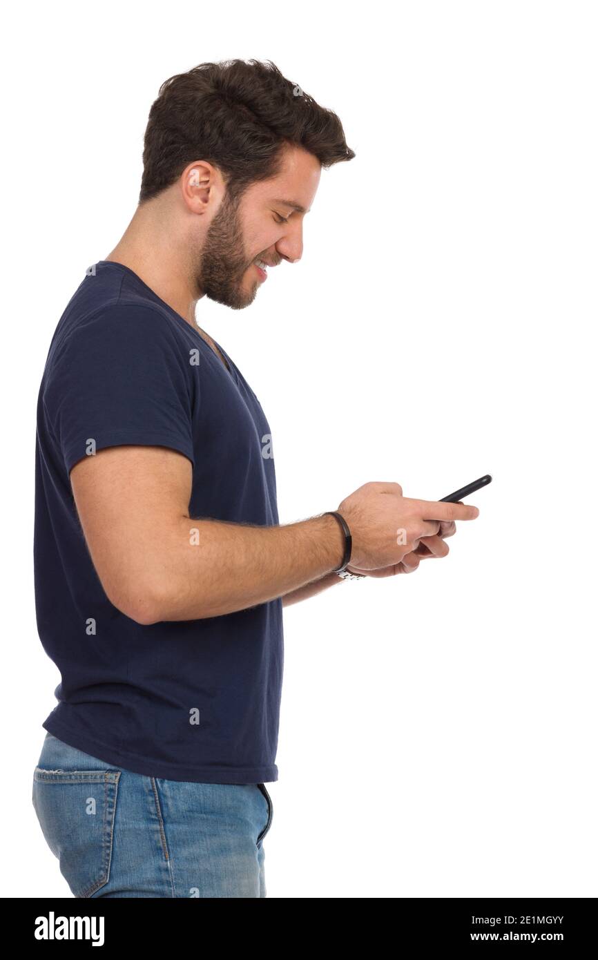 Schöner lächelnder Mann in blauem T-Shirt hält Telefon. Seitenansicht. Drei Viertel Länge Studioaufnahme isoliert auf weiß. Stockfoto