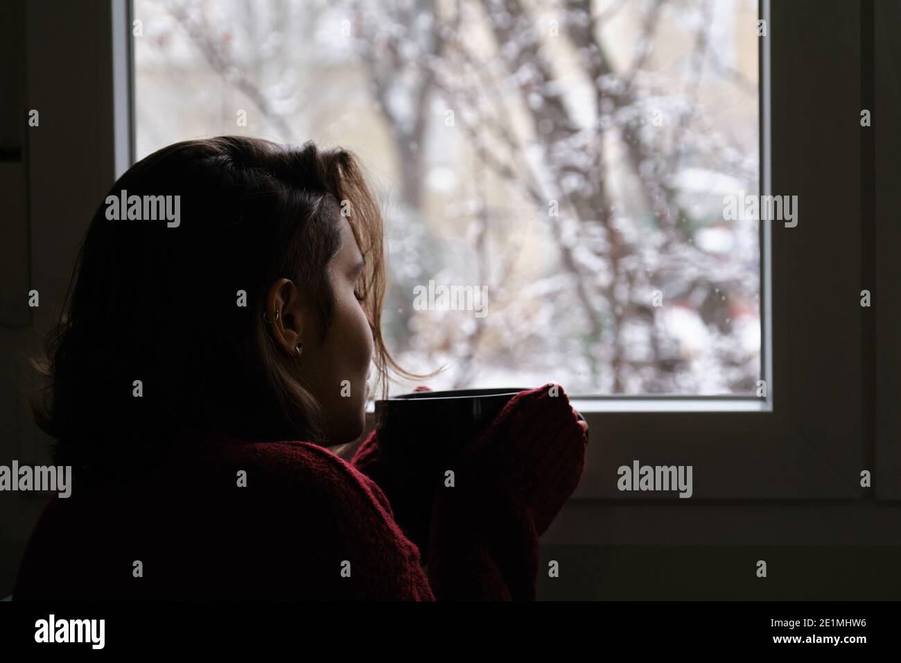 Junge Frau bläst auf heißen Kaffee oder Tee Tasse Blick aus dem Fenster in einem verschneiten Wintertag. Gemütlich zu Hause Konzept. Stockfoto