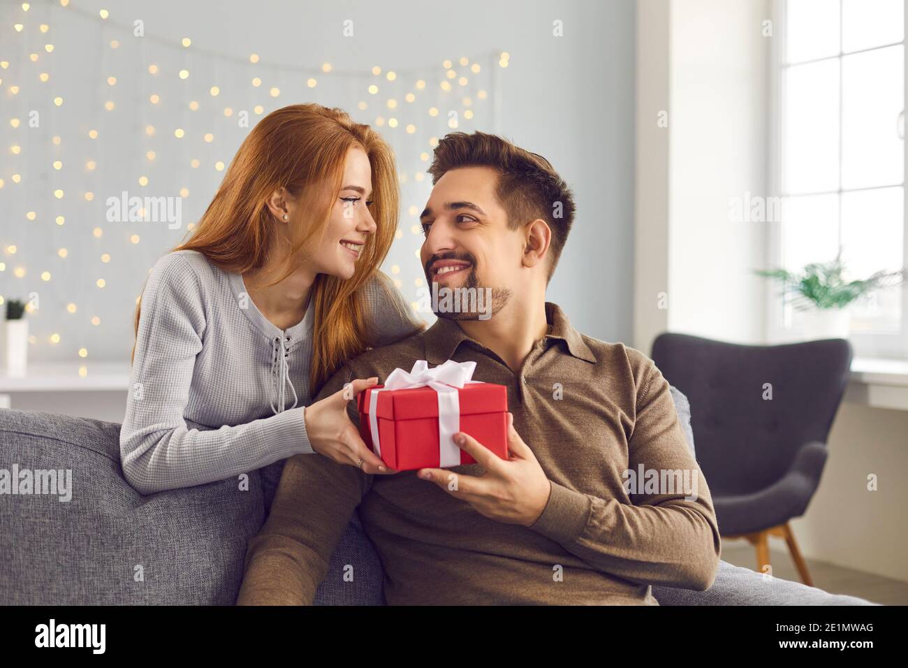 Romantische Frau steht hinter einem Mann auf einem Sofa sitzend und überreicht ihm ein Urlaubsgeschenk. Stockfoto