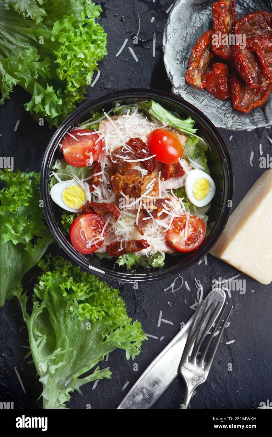Nahaufnahme von Gemüsesalat mit grünen Salatblättern, frischen und trockenen Kirschtomaten, geriebenem Parmesankäse, Eiern, Fleisch und Croutons, auf einem schwarzen Teller Stockfoto
