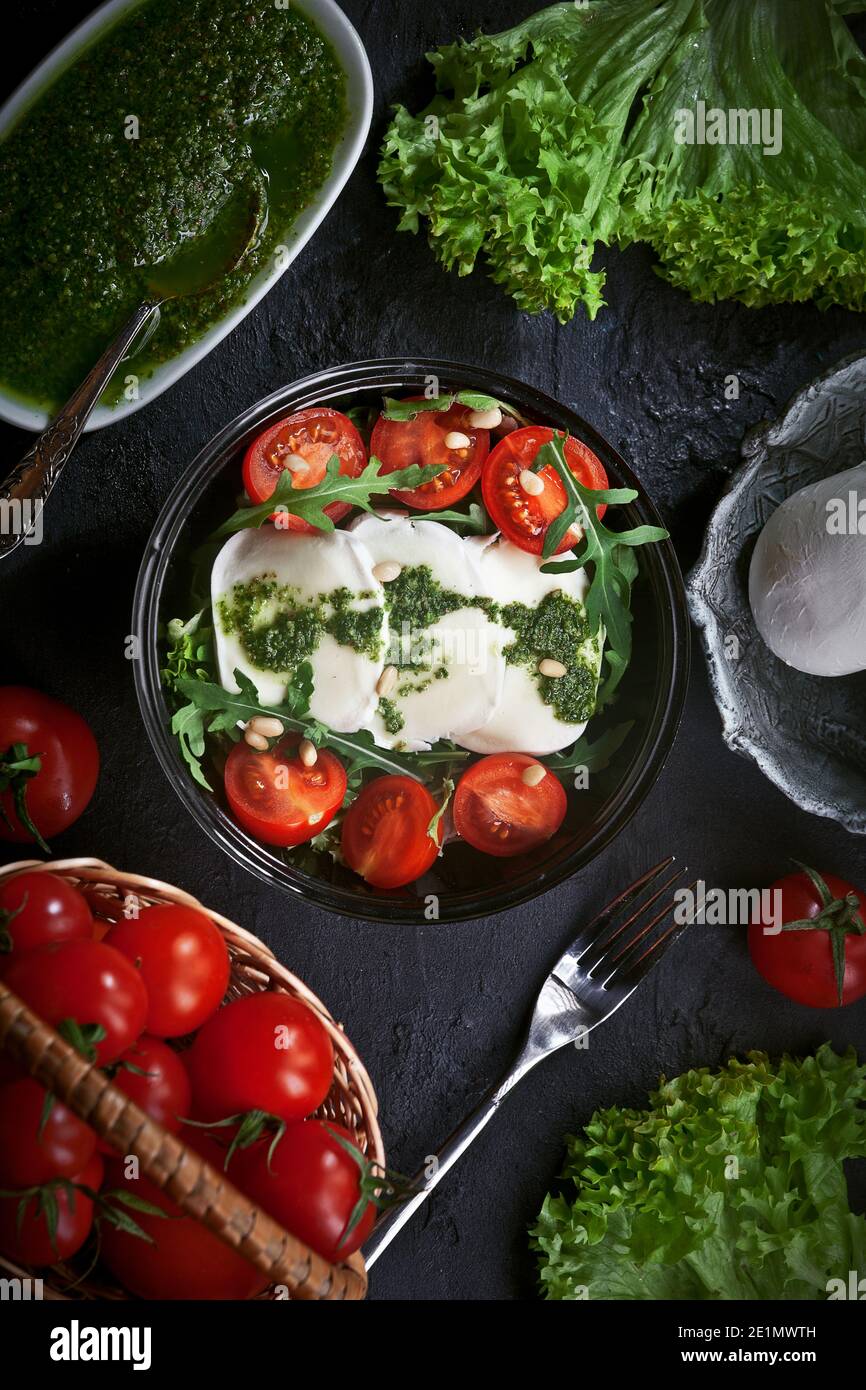 Frischer Mozzarella-Salat mit Salat und Rucola-Blättern, Pesto-Sauce, frischen Kirschtomaten und Zedernnüssen, auf schwarzem Teller auf dunklem Schiefertisch backgr Stockfoto