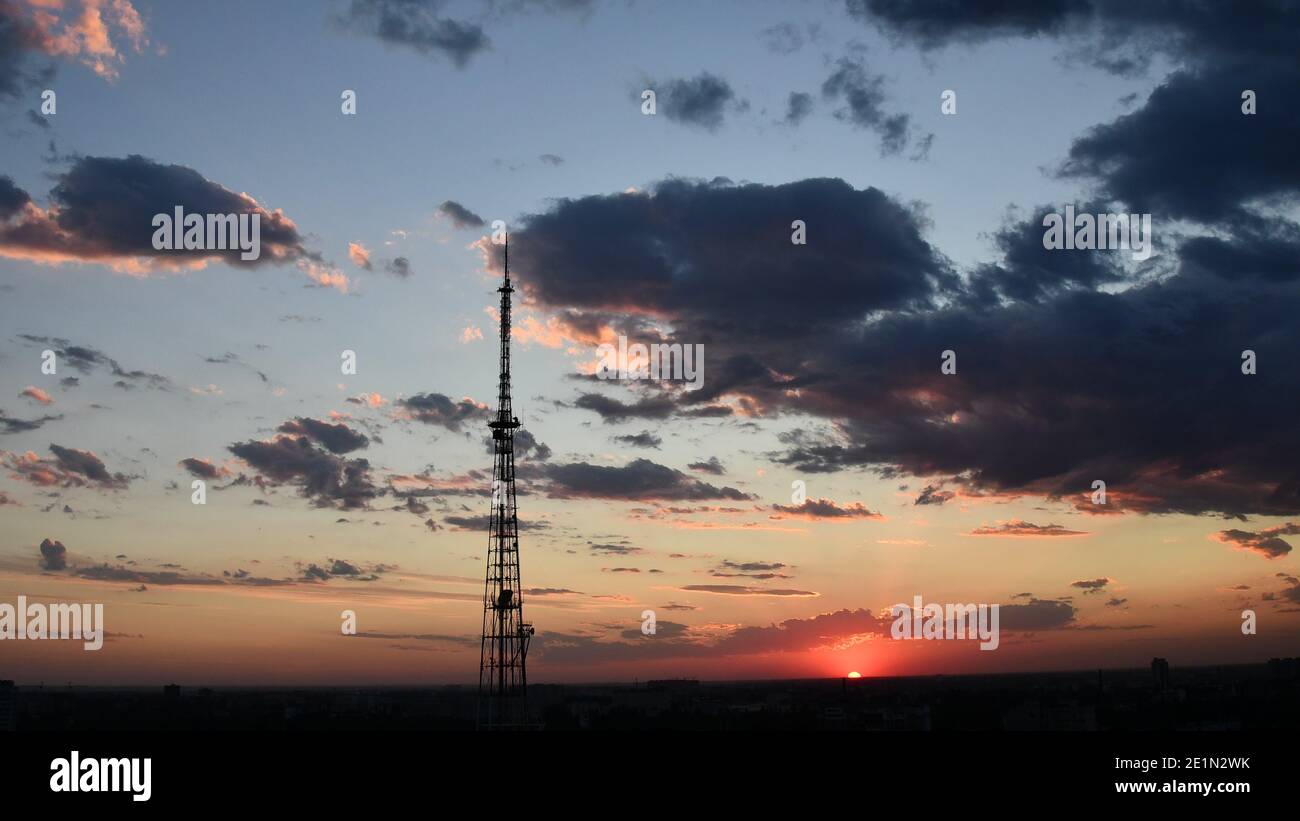 Wolkenlandschaft mit Industriekommunikationsturm und dunkelvioletten Wolken, während goldene Sonnenstrahlen durchscheinen. Rote Sonne Wolkenlandschaft des Sonnenuntergangs. Düsteres Drama Stockfoto