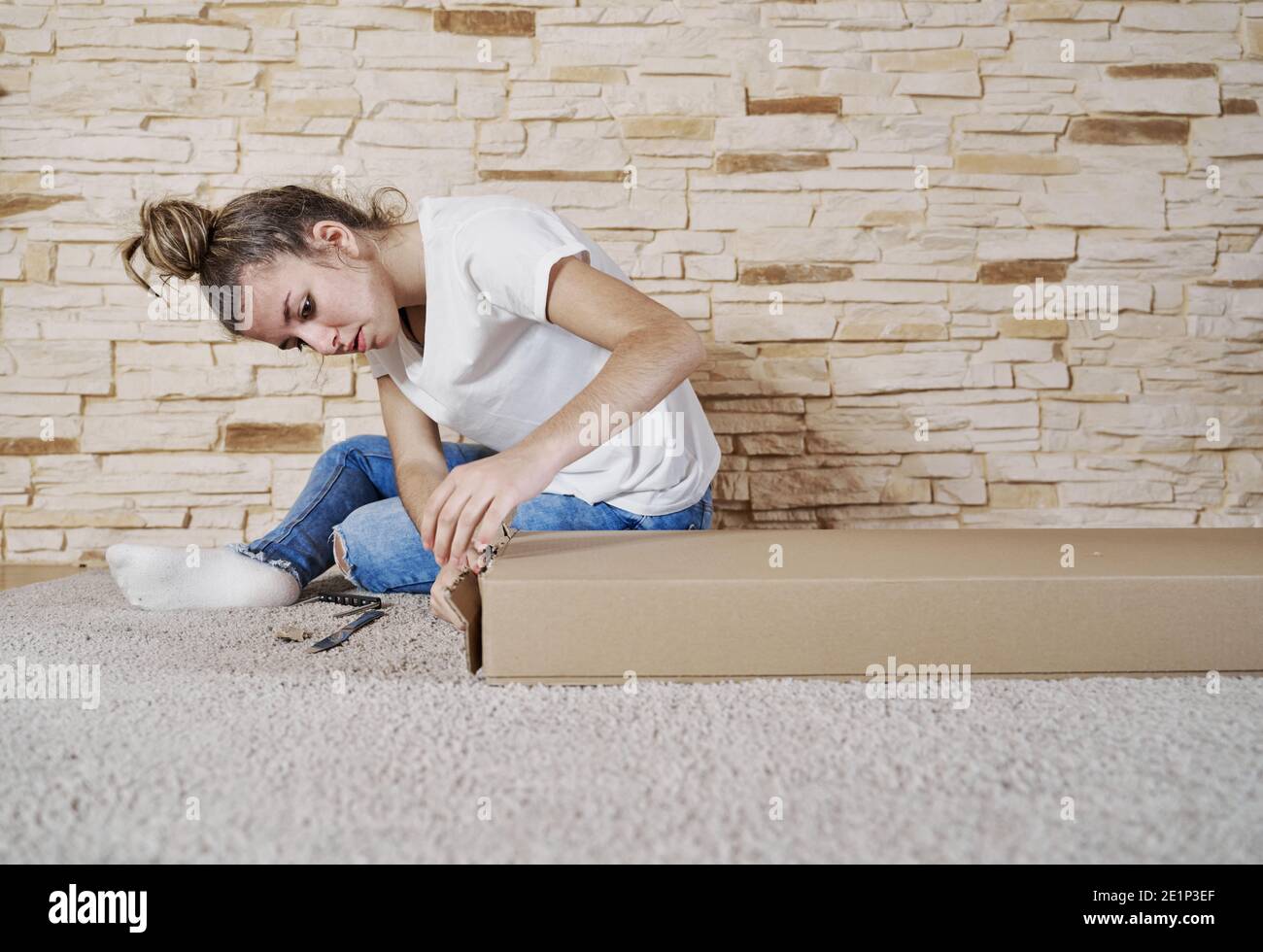 Vorderansicht des Mädchens, das eine Pappkartonschachtel mit einem Messer öffnet. Die Kiste ist auf dem Boden und die Frau sitzt. Horizontales Foto Stockfoto