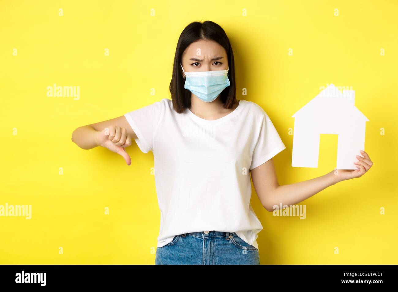 Covid-19 und Immobilienkonzept. Enttäuschte asiatische Frau in medizinische Maske, zeigt Daumen nach unten und Papierhaus Ausschnitt, stehen aufgeregt gegen gelb Stockfoto