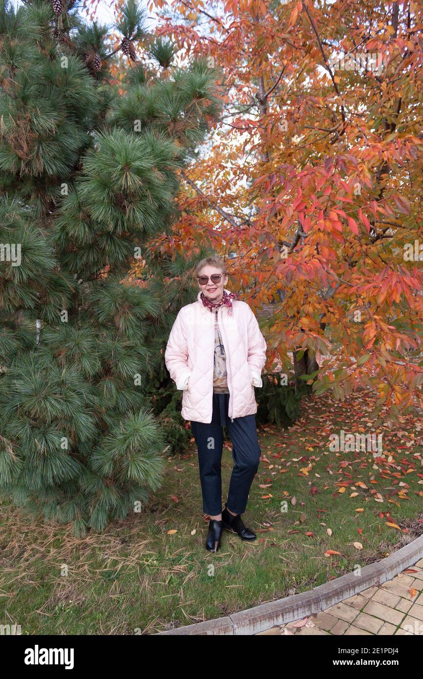 Eine reife Frau, stilvoll gekleidet, in einem Herbstpark zwischen einer grünen Kiefer und einem orange-roten Baum Stockfoto