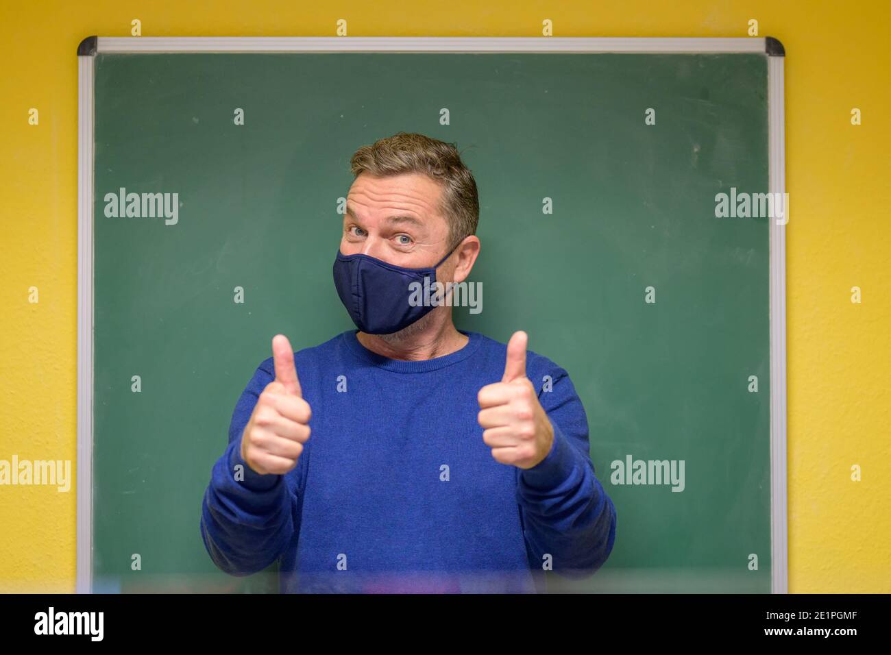 Männlicher Lehrer trägt ein Tuch Gesichtsmaske zeigt einen Erfolg Oder richtige Antwort, die einen Daumen nach oben mit beiden Händen Vor der Tafel der Klasse Stockfoto