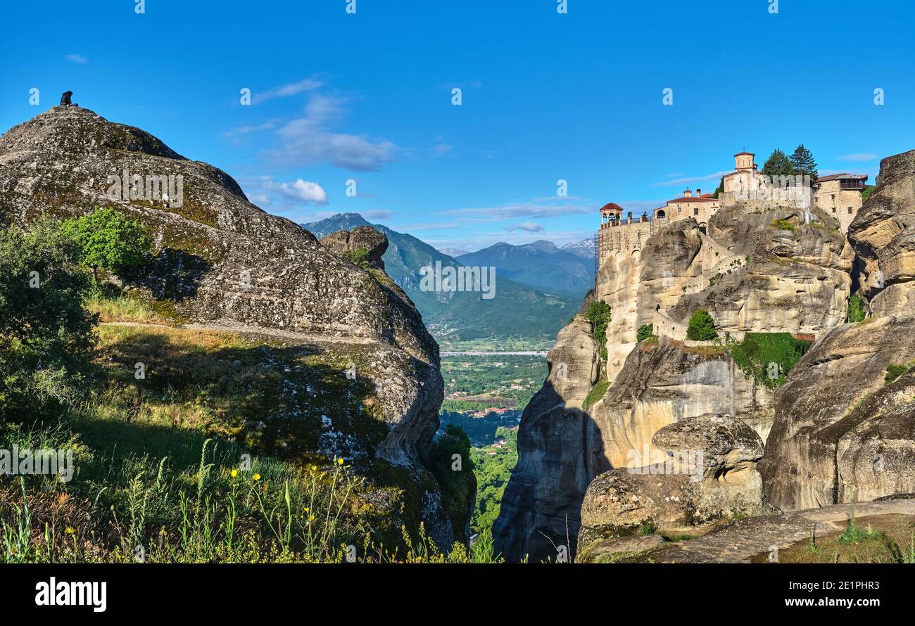 Morgenansicht des Klosters Varlaam und der Felsen. Meteora Tal, Griechenland. Nicht erkennbarer Erwachsener sitzt auf der Klippe, meditierend. UNESCO-Weltkulturerbe Stockfoto