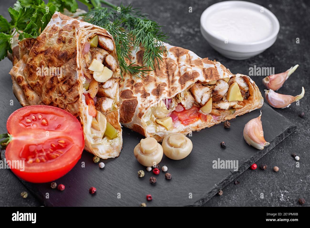 Shawarma mit Hühnerfleisch, mit Sauce, Zwiebeln, Gurken, Tomaten, Knoblauch, Kräutern und Pilzen Champignons, auf Schiefer, vor dunklem Betongrund Stockfoto