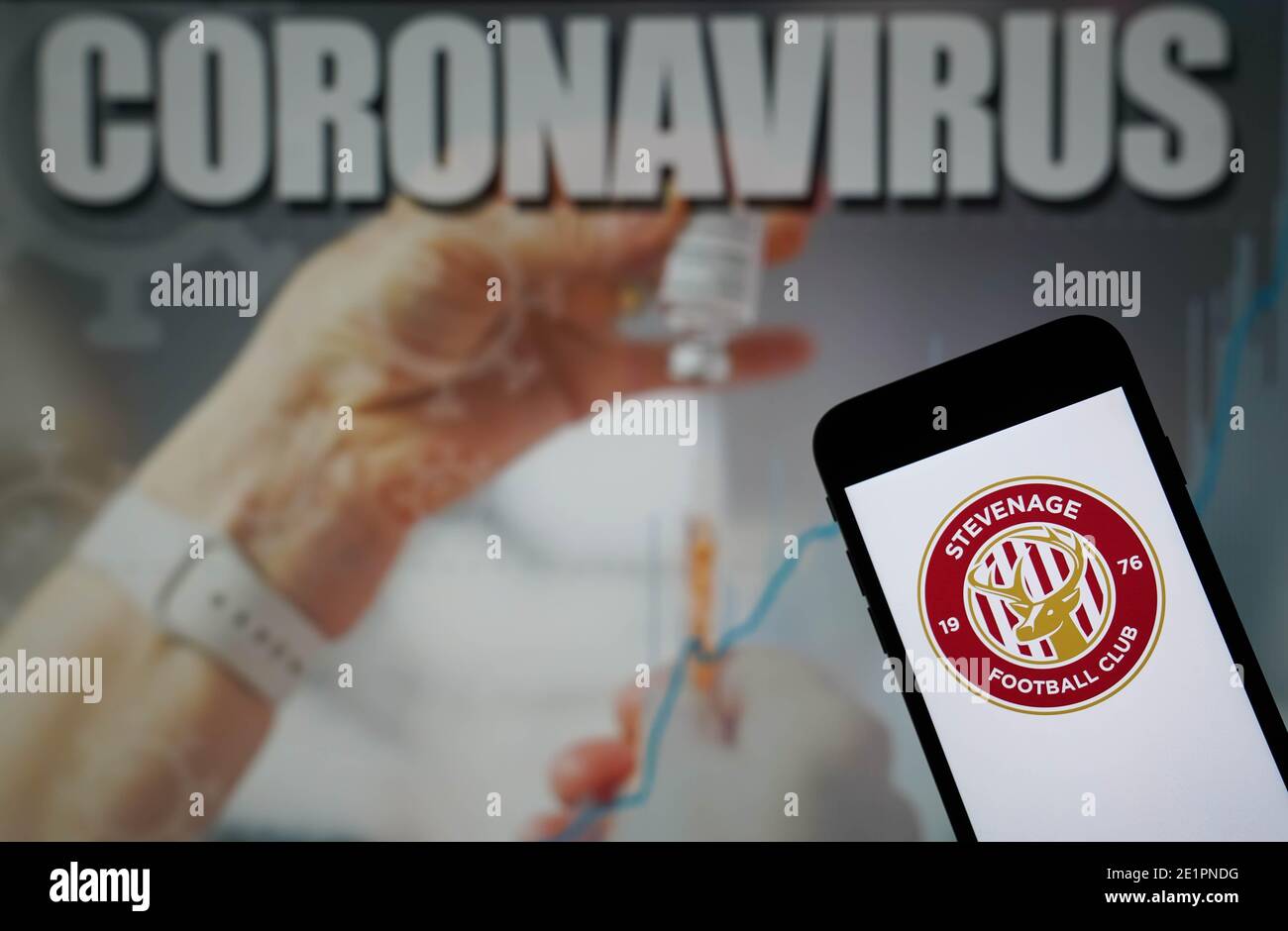 Das Stevenage Football Club-Logo wird auf einem Handy angezeigt Telefon mit einer Coronavirus-Abbildung auf einem Monitor im Hintergrund Stockfoto