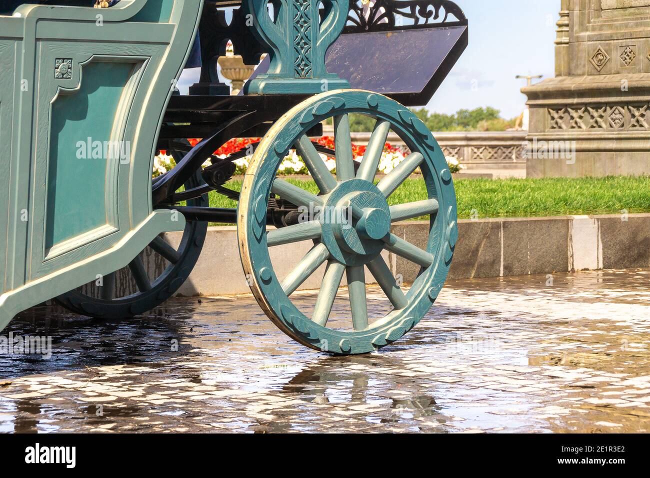 Grüner Holzwagen mit Holzrädern steht auf nassen Pflastersteinen, im Hintergrund sind Blumenbeete und Steinpodeste mit geschnitztem Oranhmen Stockfoto