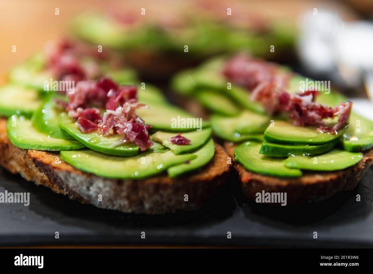 Detailreiche Aufnahme von Avocado-Toast mit iberischem Schinken auf Schiefertafeln. Sie können mehr Toasts im Hintergrund sehen, die nicht scharf sind Stockfoto