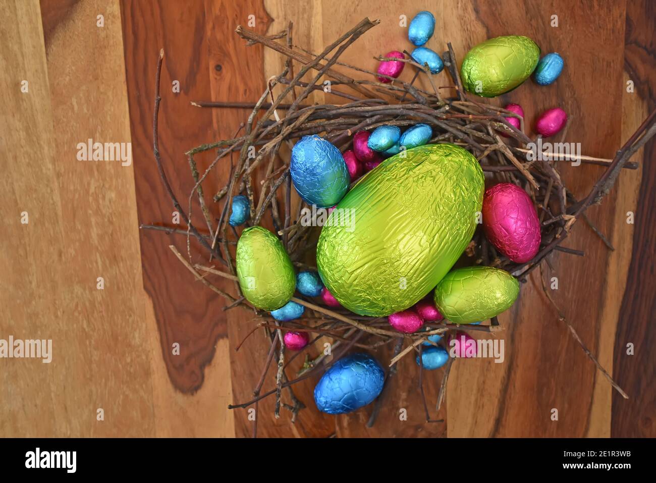 In Folie gewickelte bunte ostereier in rosa, grün, blau und gelb in einem natürlichen Nest aus Stöcken und Zweigen. In der Mitte befindet sich ein großes Ei. Stockfoto