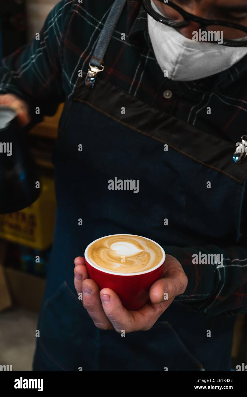 Nahaufnahme eines maskierten, nicht erkennbaren Barista, der einen Cappuccino zubereitet Eine rote Tasse Stockfoto