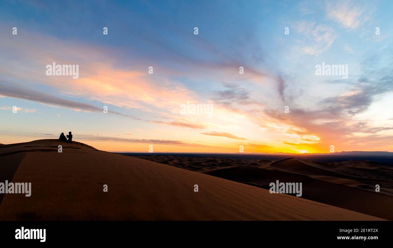(Selektiver Fokus) Silhouette eines Paares, das auf den Sanddünen der Merzouga-Wüste bei einem atemberaubenden Sonnenuntergang sitzt. Merzouga, Marokko. Stockfoto