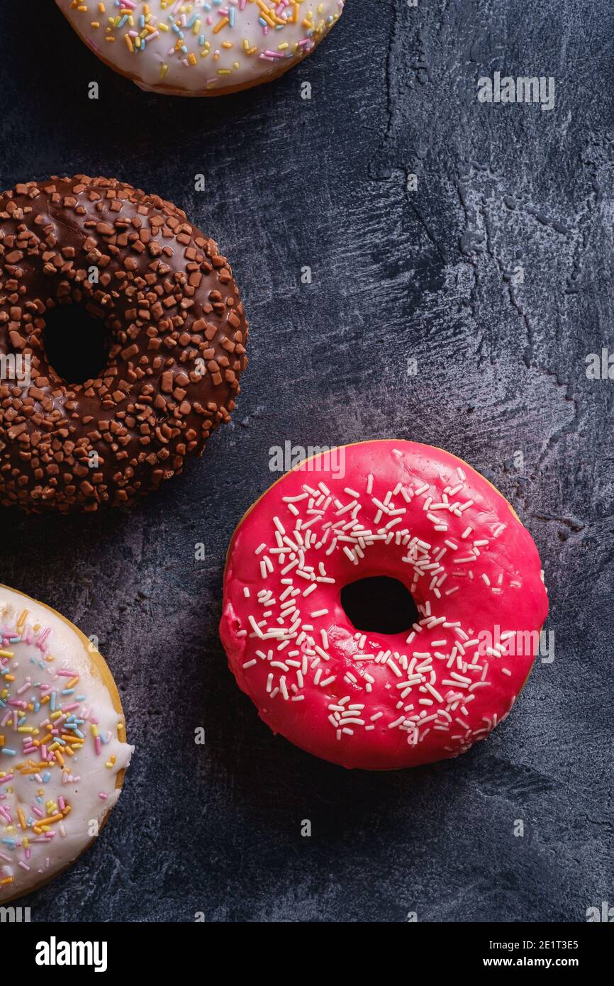 Donuts aus Schokolade, Rosa und Vanille mit Streuseln, süße glasierte Dessertspeisen auf dunklem Beton-Strukturhintergrund, Draufsicht Stockfoto