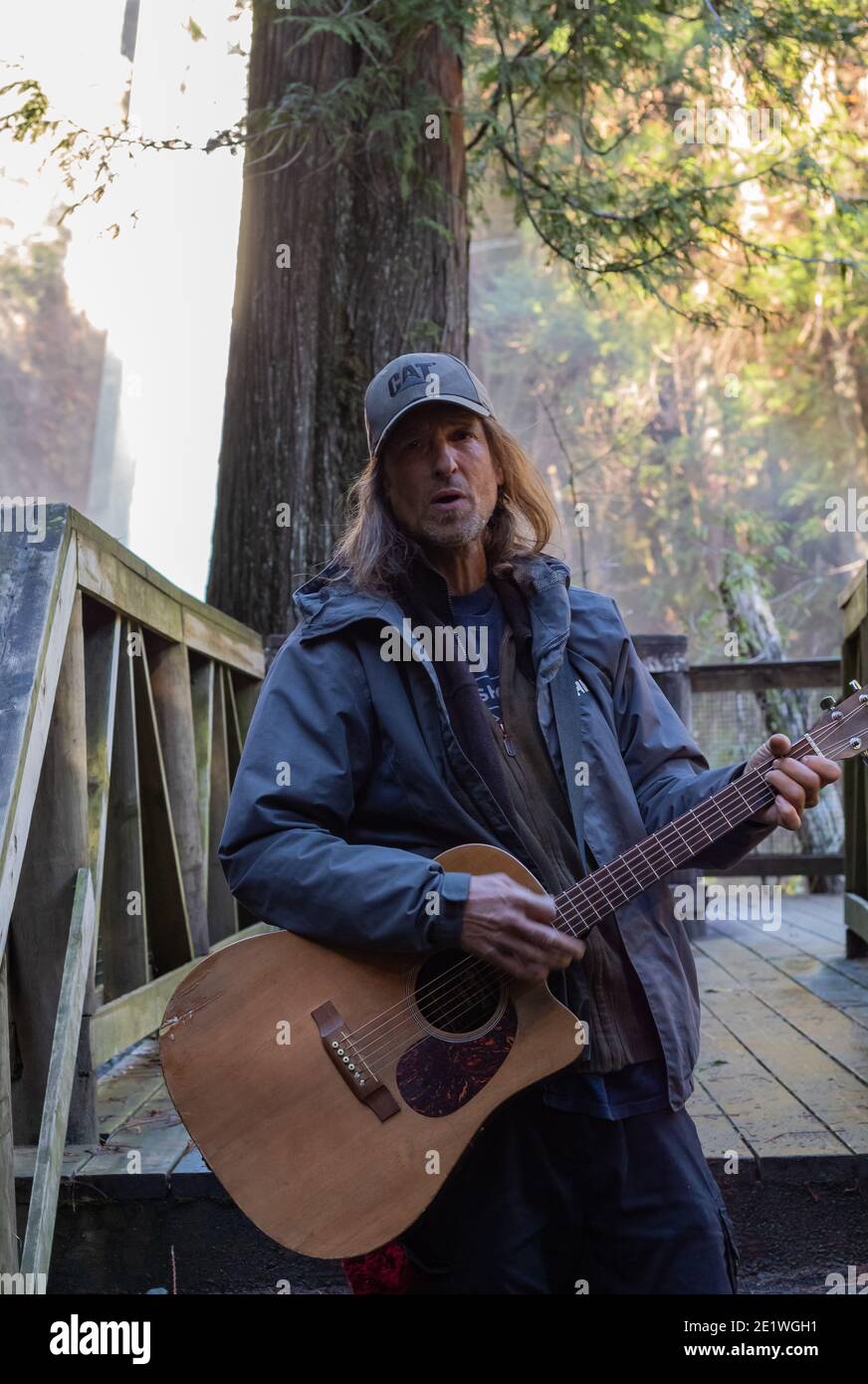 Ein Mann mittleren Alters, der einen Hut trägt und Gitarre spielt Bei der Aussichtspunktbrücke im Park von West Vancouver, BC Stockfoto