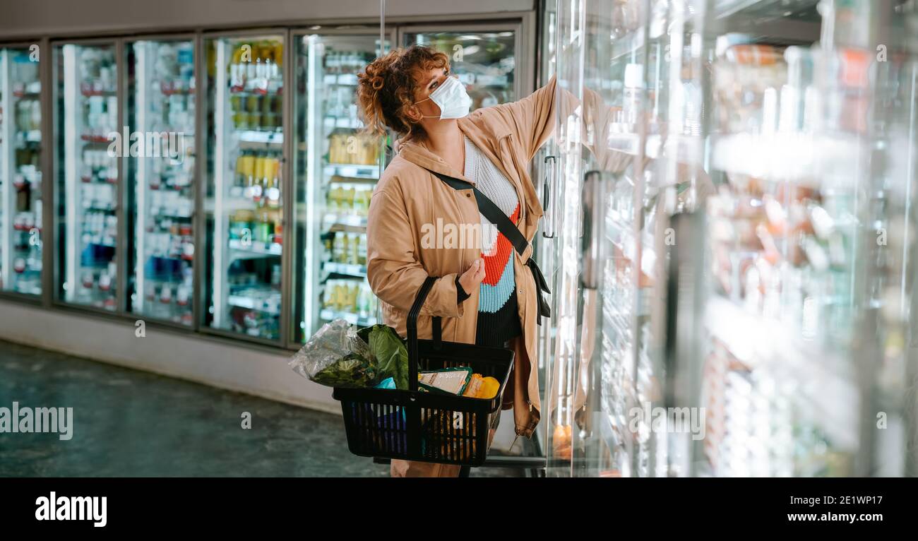 Frau, die Produkte aus dem Regallager abholt. Frau mit Gesichtsmaske einkaufen Lebensmittel im Supermarkt. Stockfoto