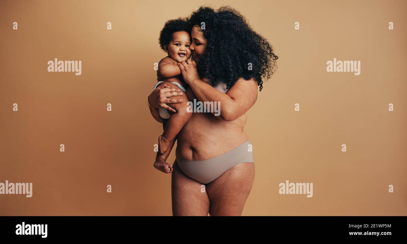 Plus Größe Frau mit ihrem Baby. Glückliche Mama trägt ihr Kleinkind im Studio. Echter Frauenkörper nach der Geburt des Kindes. Stockfoto