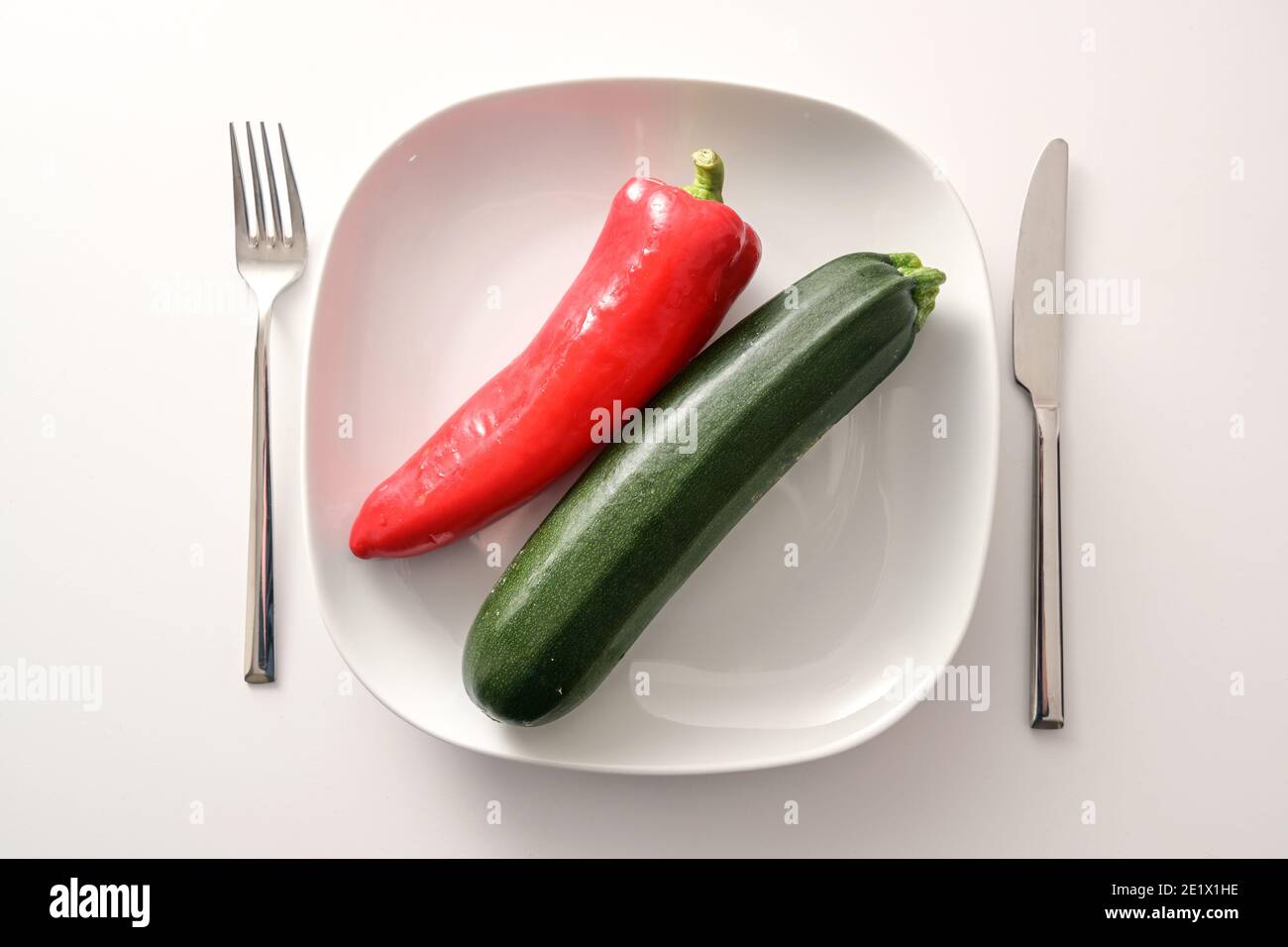 Roher roter, spitzer Pfeffer und Zucchini auf weißem Teller und Besteck auf hellem Hintergrund, gesunde Ernährung mit mediterranem Gemüse, um Gewicht zu verlieren Stockfoto