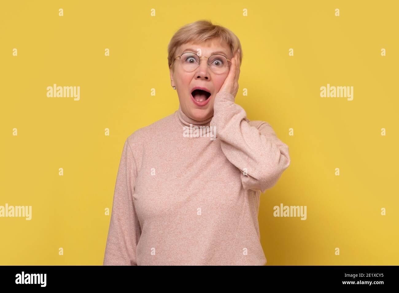Erstaunt reife Frau in Brille mit geöffnetem Mund fühlt sich betäubt nach schockierend neu. Studio auf gelber Wand. Stockfoto
