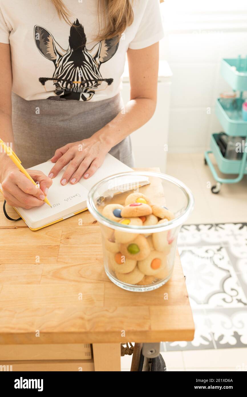 Fokussierte Frau Hände mit gepflegten Nägeln Schreiben ein Plätzchen Rezept mit einem gelben Bleistift in der Nähe eines transparenten Glas voller bunte Kekse. Stockfoto