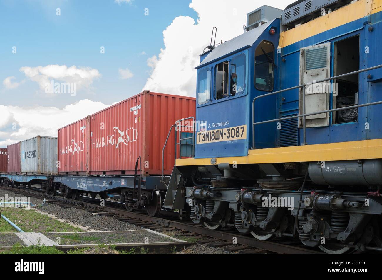 An einem sonnigen Sommertag steht eine Lokomotive mit Fahrer im Fahrerhaus als Teil eines Güterzuges mit Containern auf der Eisenbahnstrecke. Stockfoto