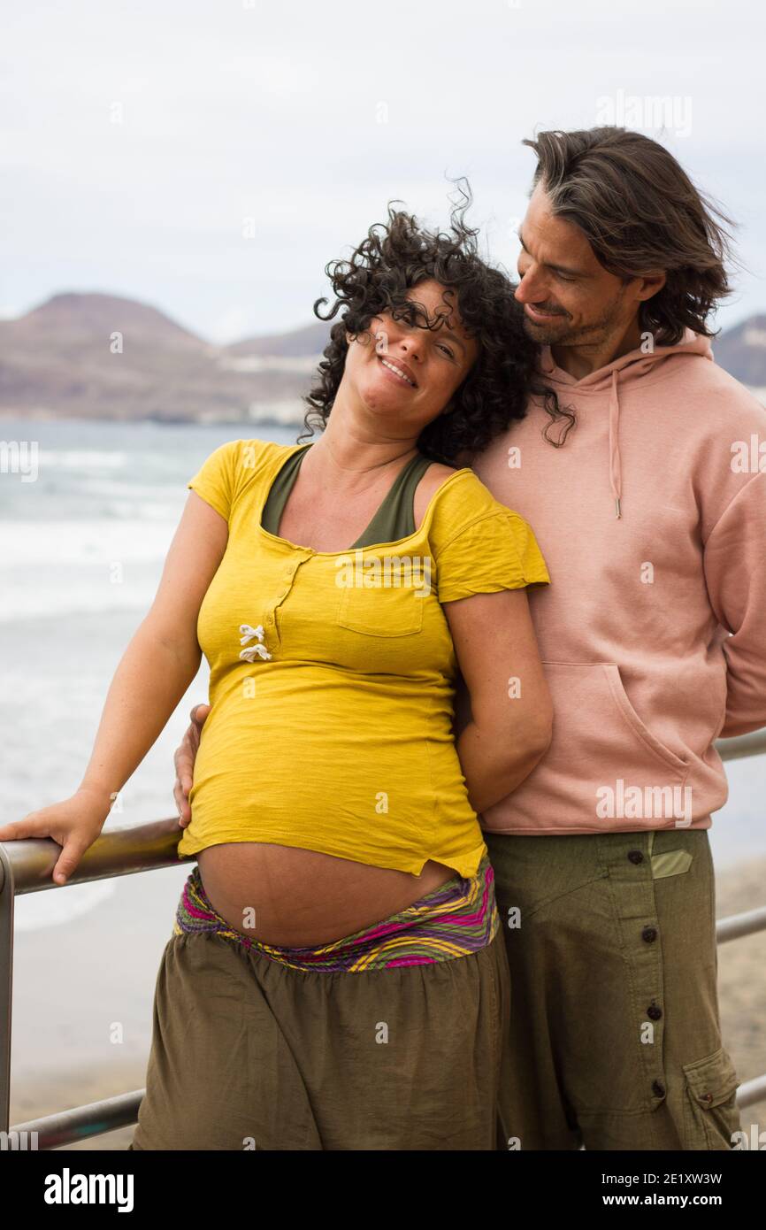 Glückliches Paar erwartet Baby auf Strandspaziergang. Schwangere Frau auf gelbem Hemd und lächelnder Vater, der sie an einem windigen Tag in Las Palmas, Spanien, ansieht Stockfoto