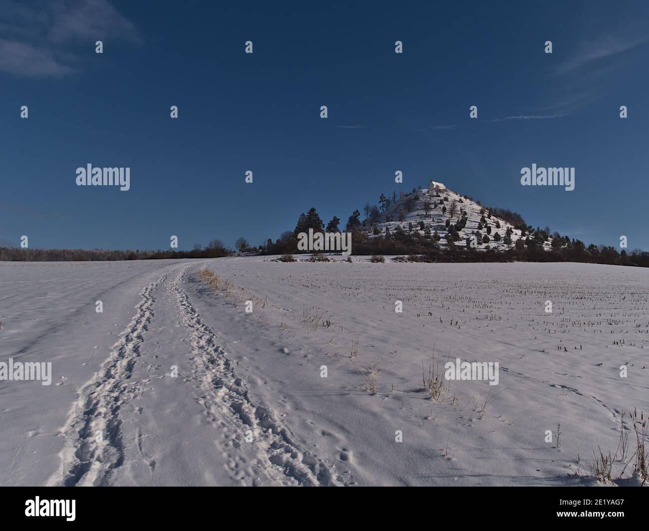 Schöne Winterlandschaft mit Spuren auf einem schneebedeckten Feld und butte Hügel Kornbühl mit historischer Kapelle Salmendinger Kapelle auf der Spitze in Deutschland. Stockfoto