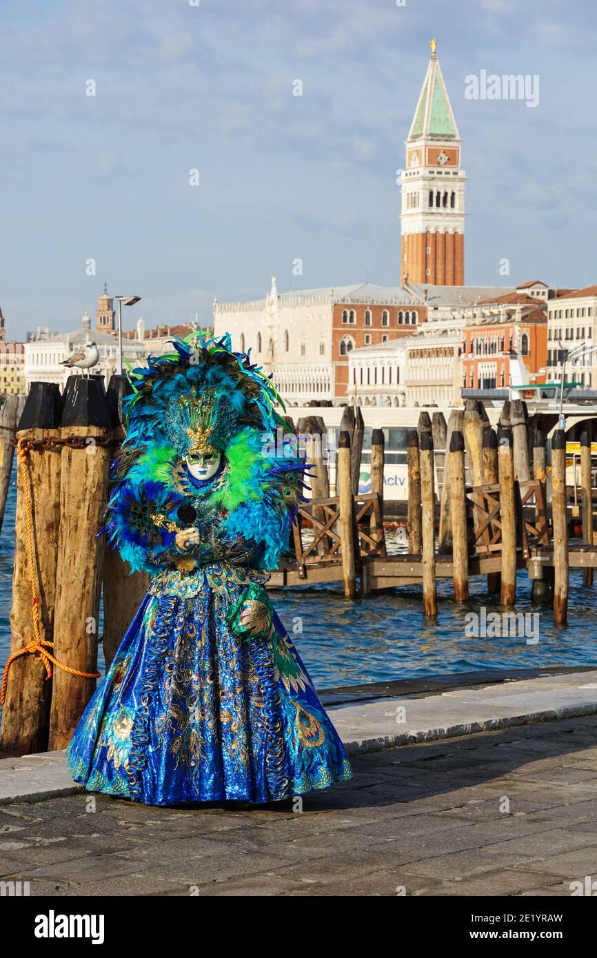 Frau gekleidet in traditionell dekoriertem Kostüm mit Federn und bemalter Maske während des Karnevals in Venedig mit dem Markusplatz Campanile hinter Venedig Italien Stockfoto