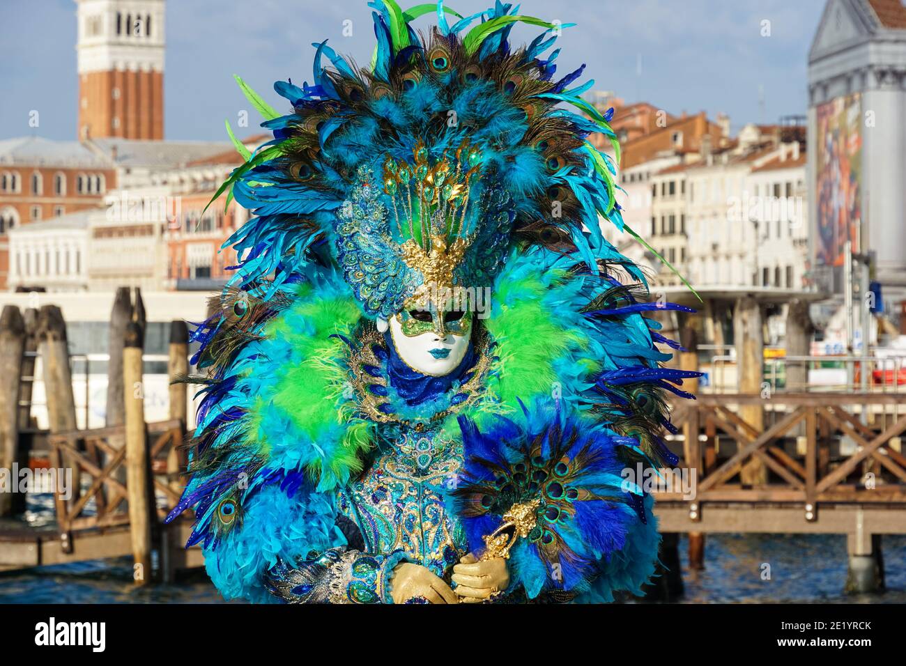 Frau gekleidet in traditionell dekoriertem Kostüm mit Federn und bemalter Maske während des Karnevals in Venedig mit dem Markusplatz Campanile hinter Venedig Italien Stockfoto