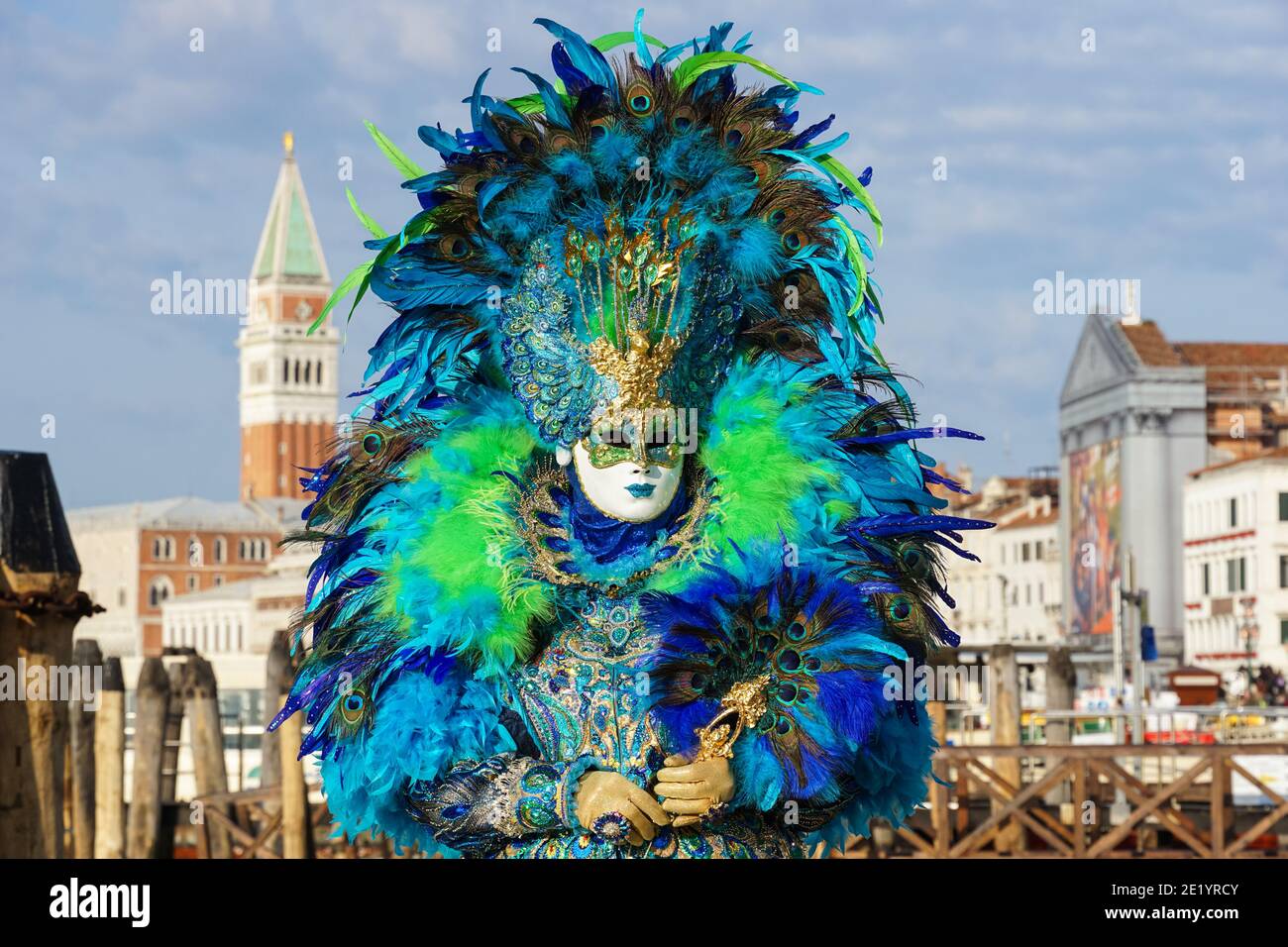 Frau gekleidet in traditionell dekoriertem Kostüm mit Federn und bemalter Maske während des Karnevals in Venedig mit dem Markusplatz Campanile hinter Venedig Italien Stockfoto